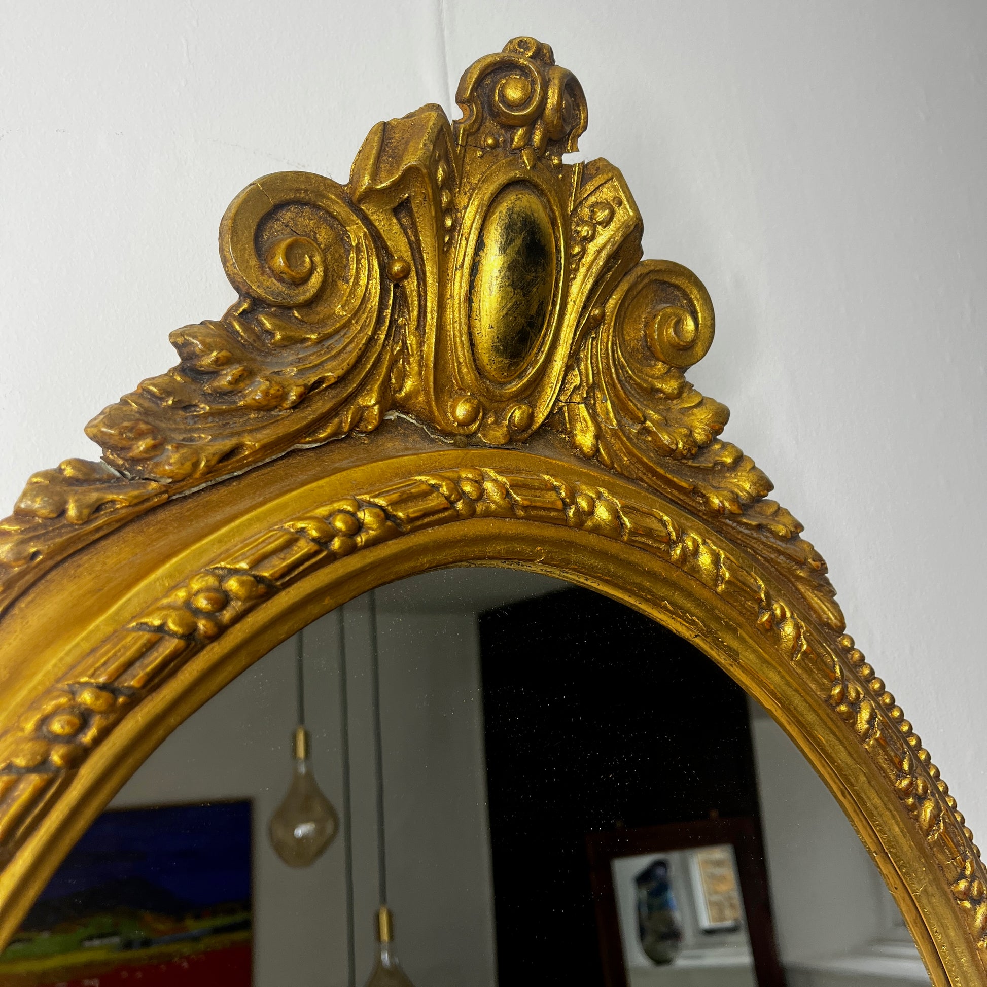 Oval gilded mirror with an ornate decorative frame featuring intricate detailing. The gold finish has a softly aged patina, giving the mirror an elegant, vintage appearance.