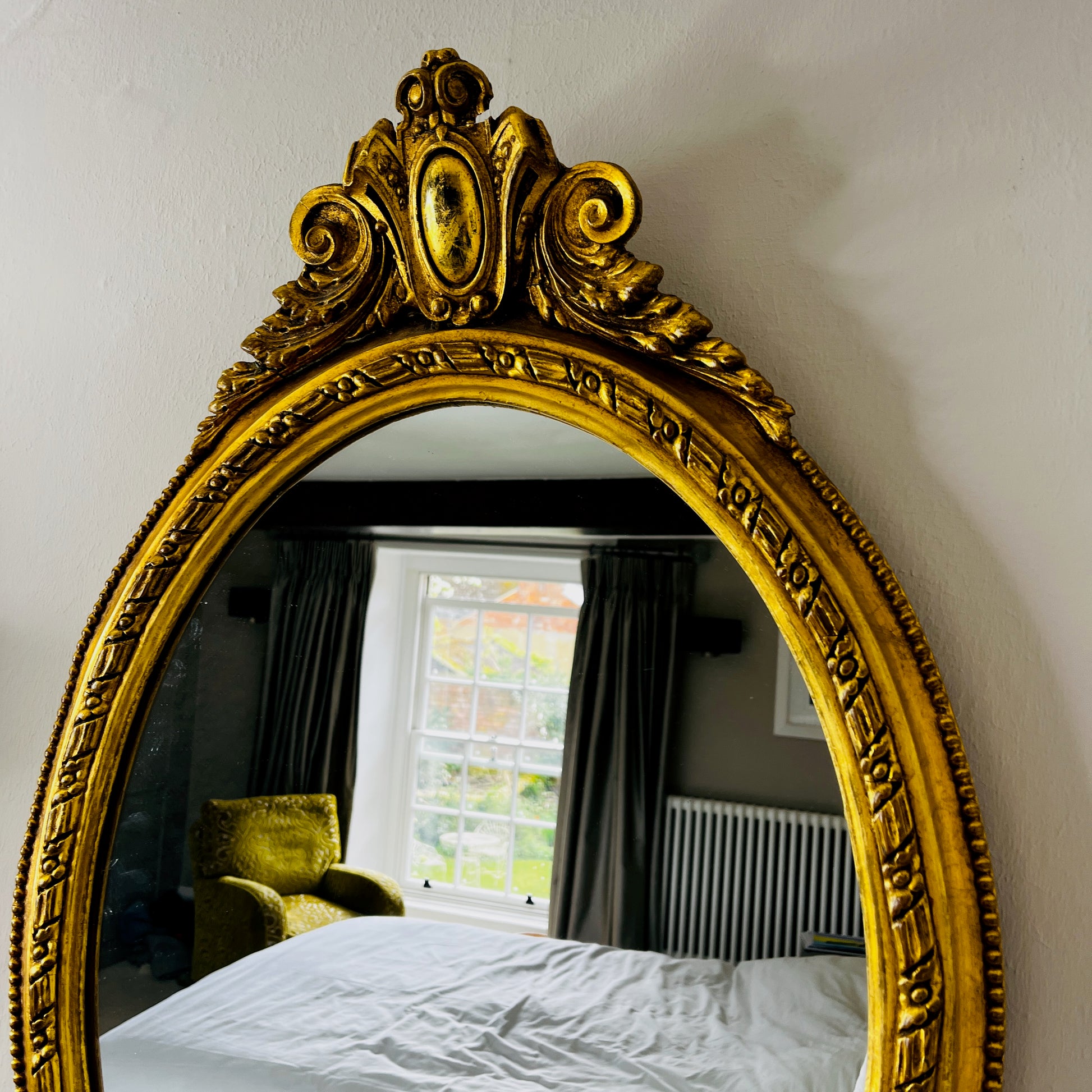 Oval gilded mirror with an ornate decorative frame featuring intricate detailing. The gold finish has a softly aged patina, giving the mirror an elegant, vintage appearance.