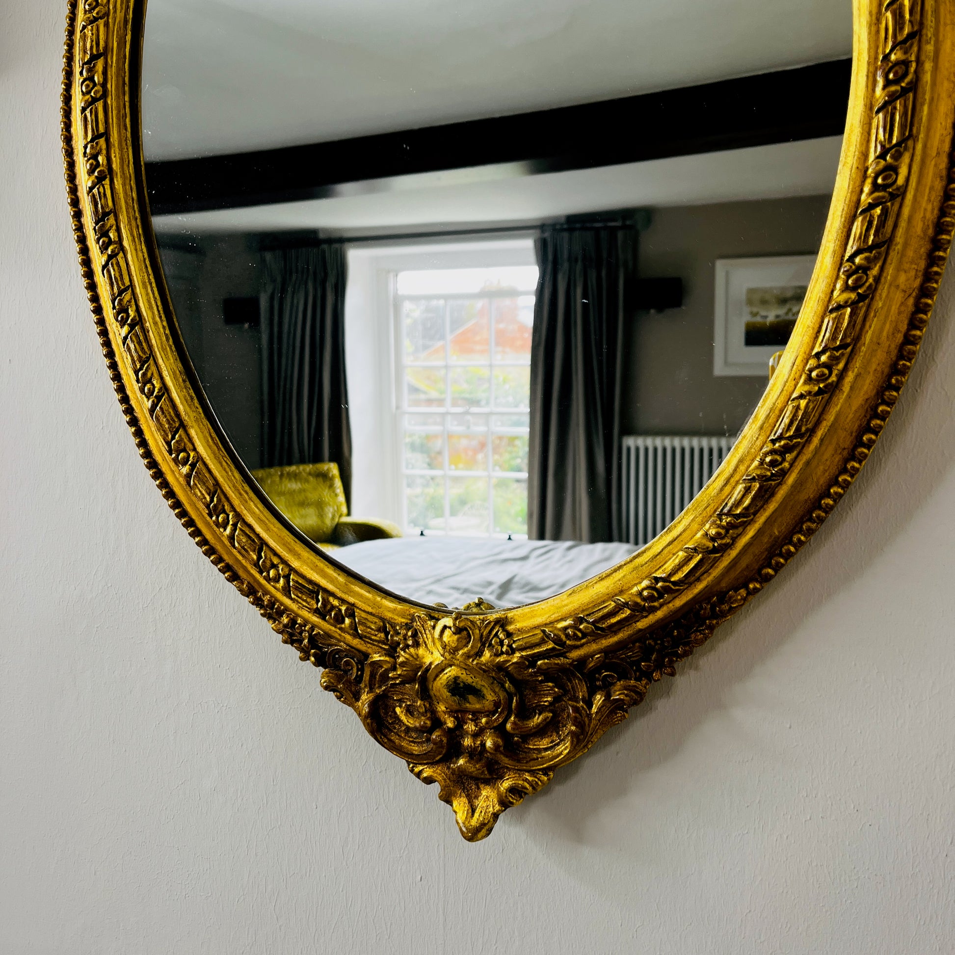 Oval gilded mirror with an ornate decorative frame featuring intricate detailing. The gold finish has a softly aged patina, giving the mirror an elegant, vintage appearance.