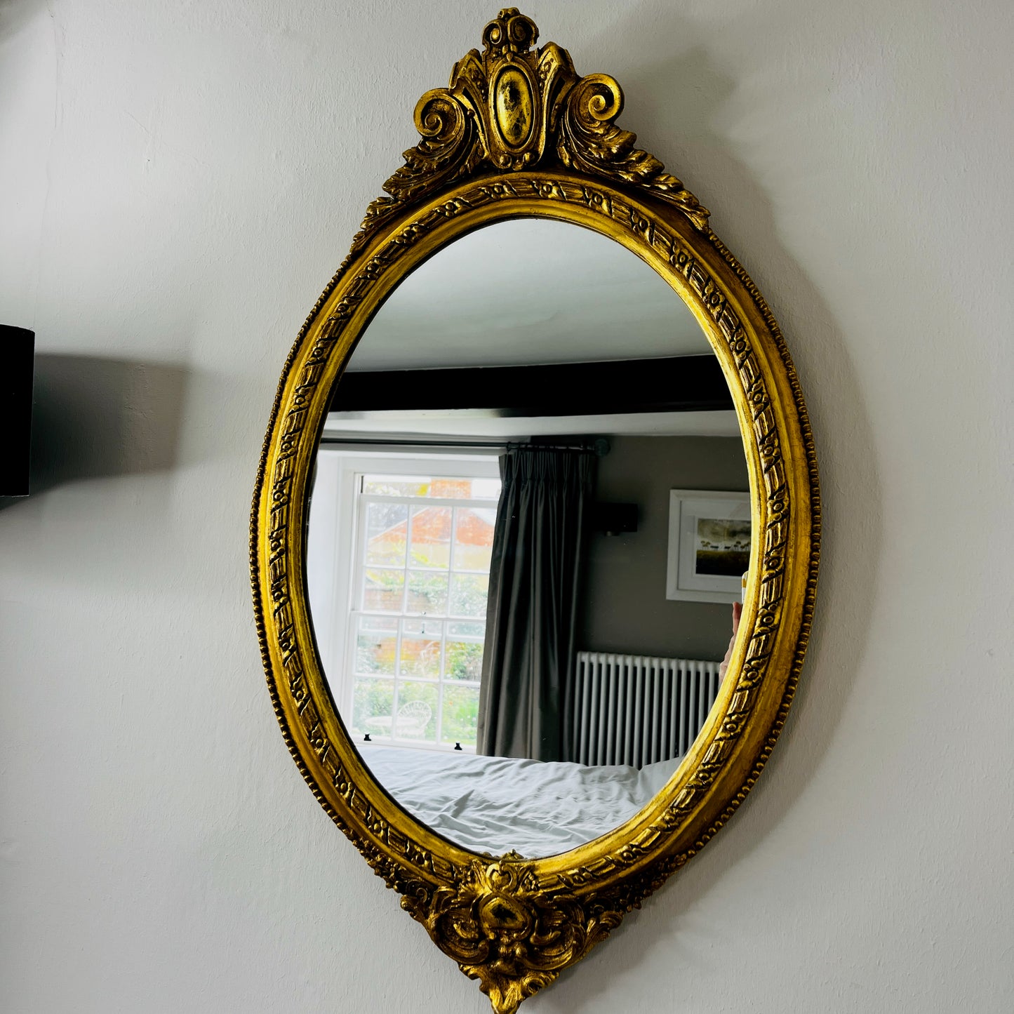Oval gilded mirror with an ornate decorative frame featuring intricate detailing. The gold finish has a softly aged patina, giving the mirror an elegant, vintage appearance.