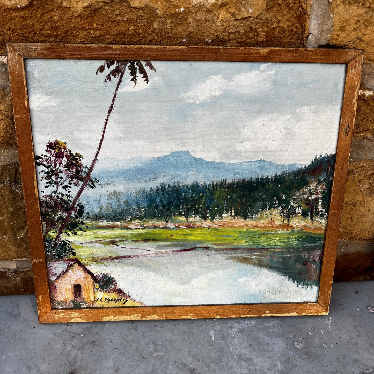 Framed vintage oil on board painting of a mountain lake scene, depicting tranquil water surrounded by rugged peaks and soft atmospheric light.
