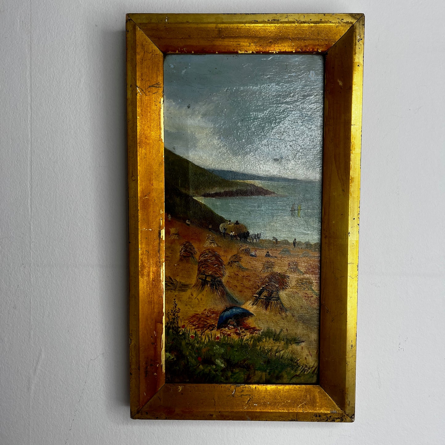 A vintage oil painting on canvas depicting a tranquil coastal scene with blue water, a sandy shoreline, and distant cliffs or headland beneath a softly painted sky. The atmospheric landscape is presented in an ornate gold-coloured frame, adding a classic, elegant touch to the artwork