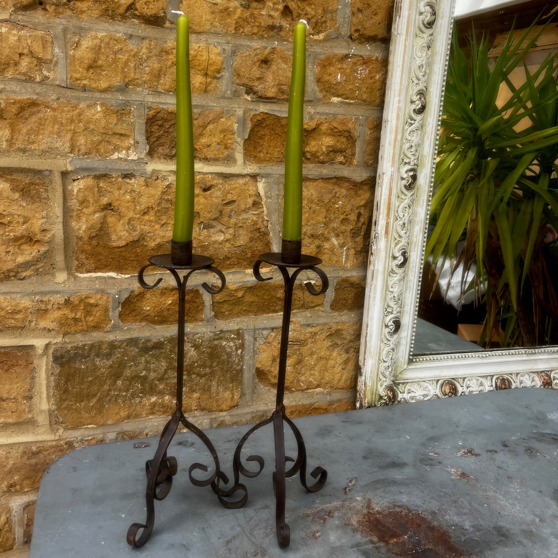 A pair of rustic black metal French-style candle holders with an aged, weathered finish. Each features an elegant turned stem, decorative detailing, and a sturdy tripod base
