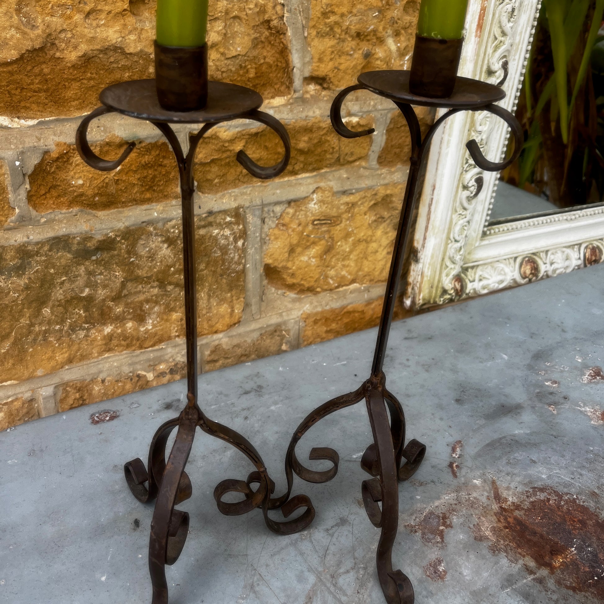 A pair of rustic black metal French-style candle holders with an aged, weathered finish. Each features an elegant turned stem, decorative detailing, and a sturdy tripod base