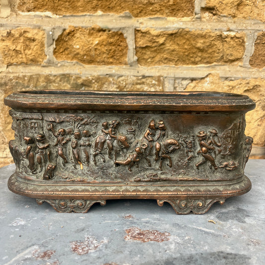 A vintage terracotta jardinière with a rich, earthy finish, featuring an intricately moulded frieze scene of classical figures and animals encircling its exterior. The cylindrical planter has a slightly weathered texture, exuding rustic elegance