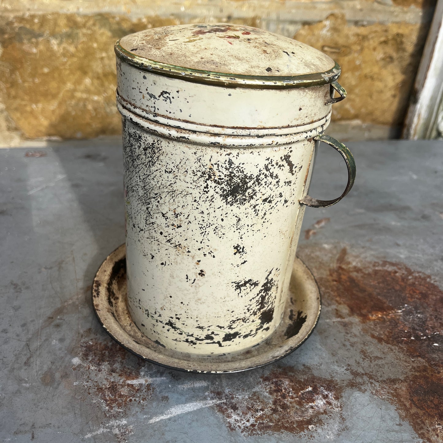 A vintage distressed metal canister with a colorful floral hand-painted design (toleware), sitting on a matching metal saucer.