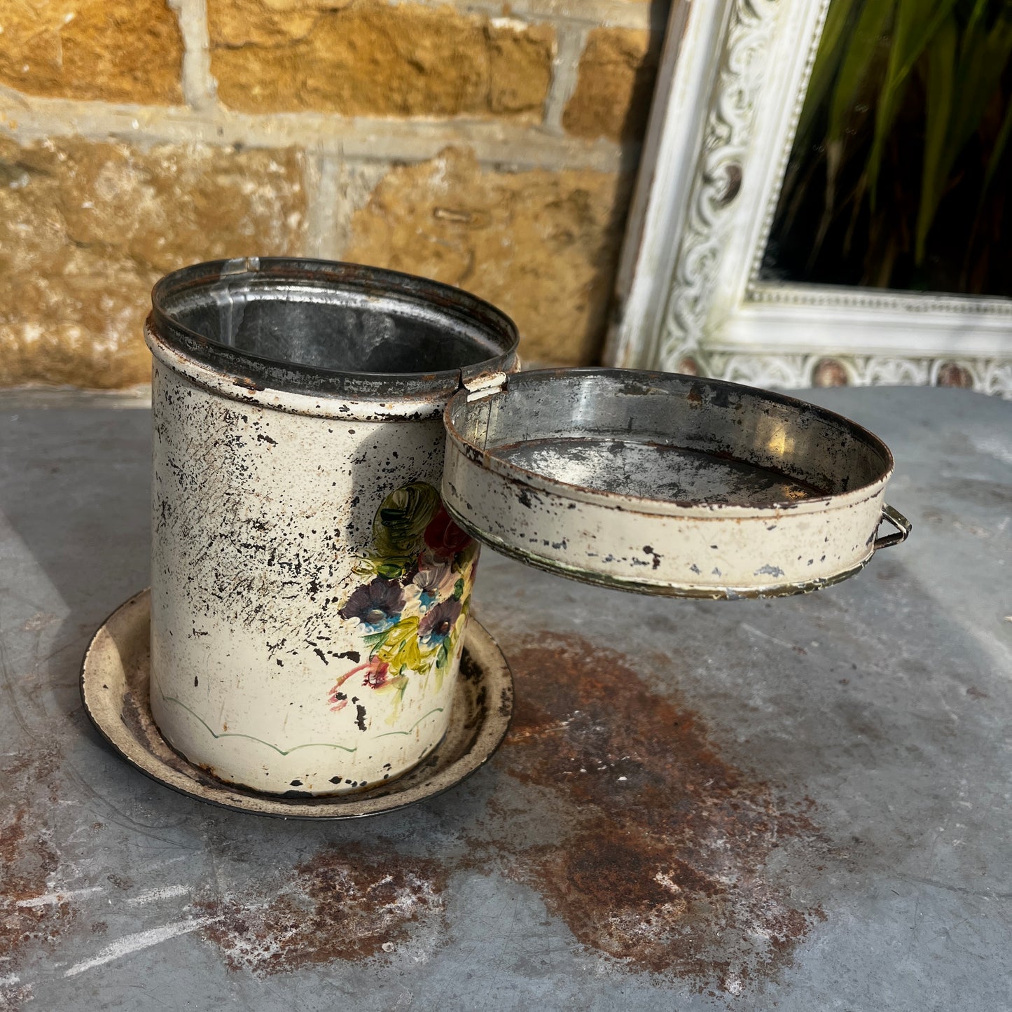 A vintage distressed metal canister with a colorful floral hand-painted design (toleware), sitting on a matching metal saucer.