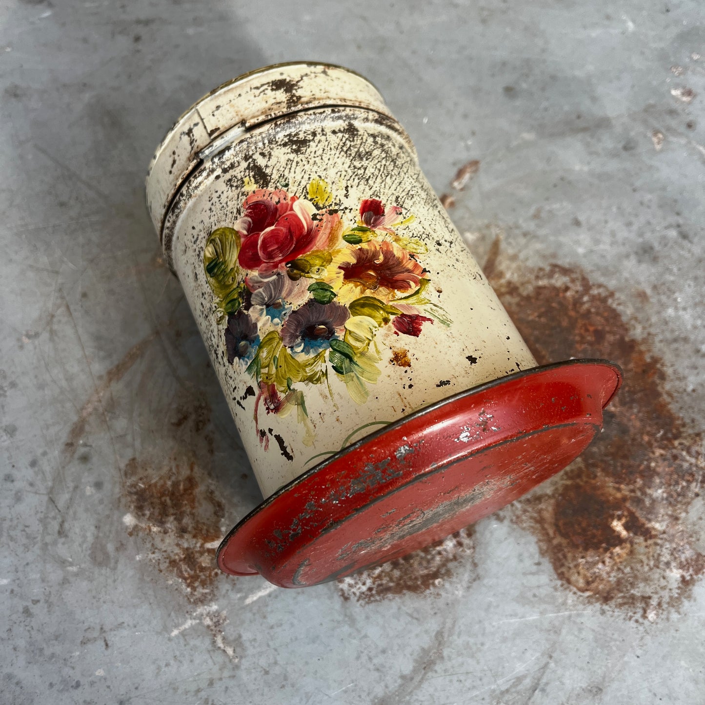 A vintage distressed metal canister with a colorful floral hand-painted design (toleware), sitting on a matching metal saucer.