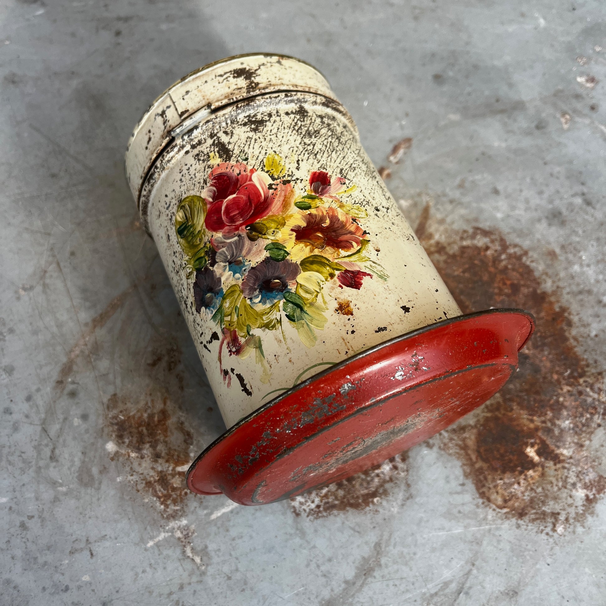 A vintage distressed metal canister with a colorful floral hand-painted design (toleware), sitting on a matching metal saucer.