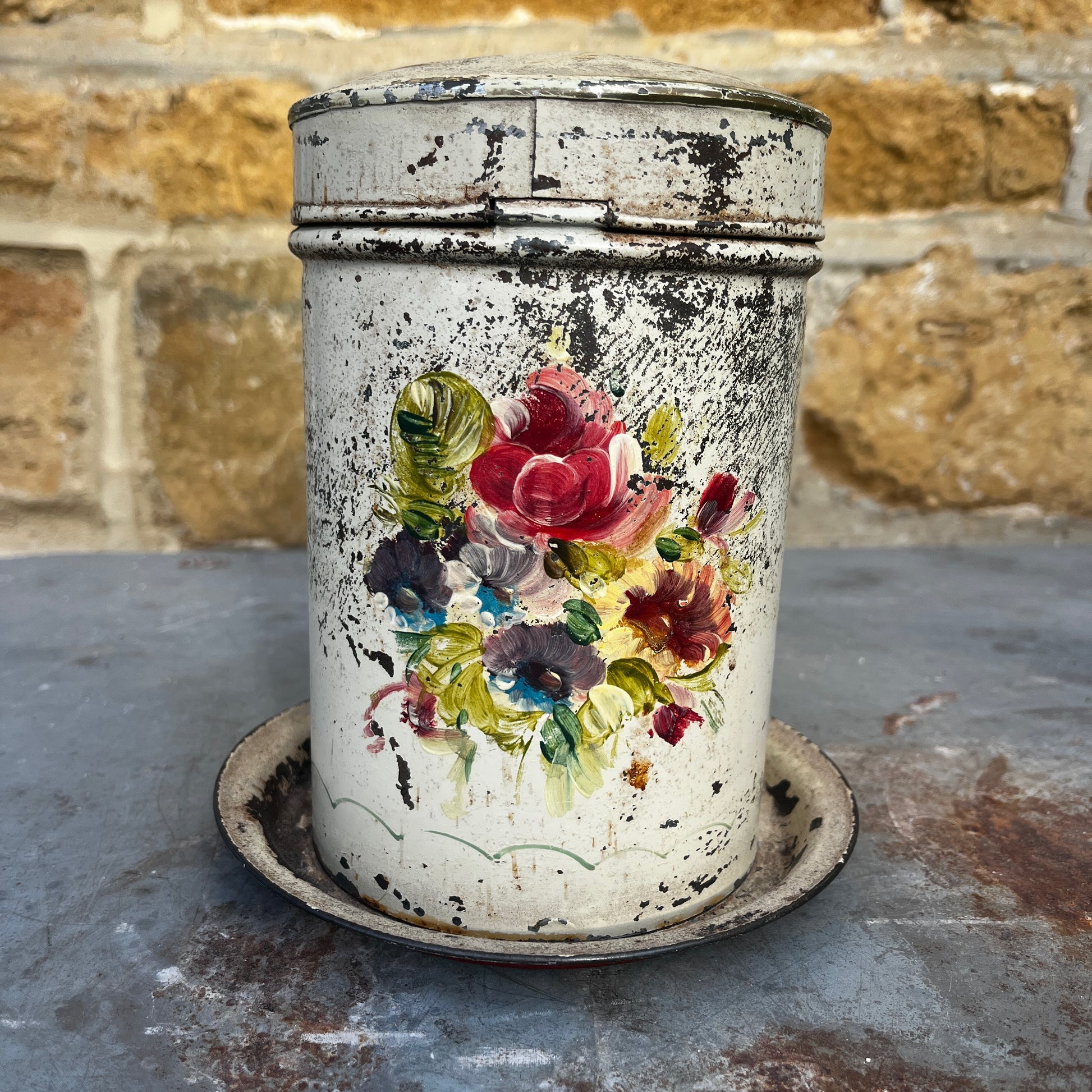 A vintage distressed metal canister with a colorful floral hand-painted design (toleware), sitting on a matching metal saucer.