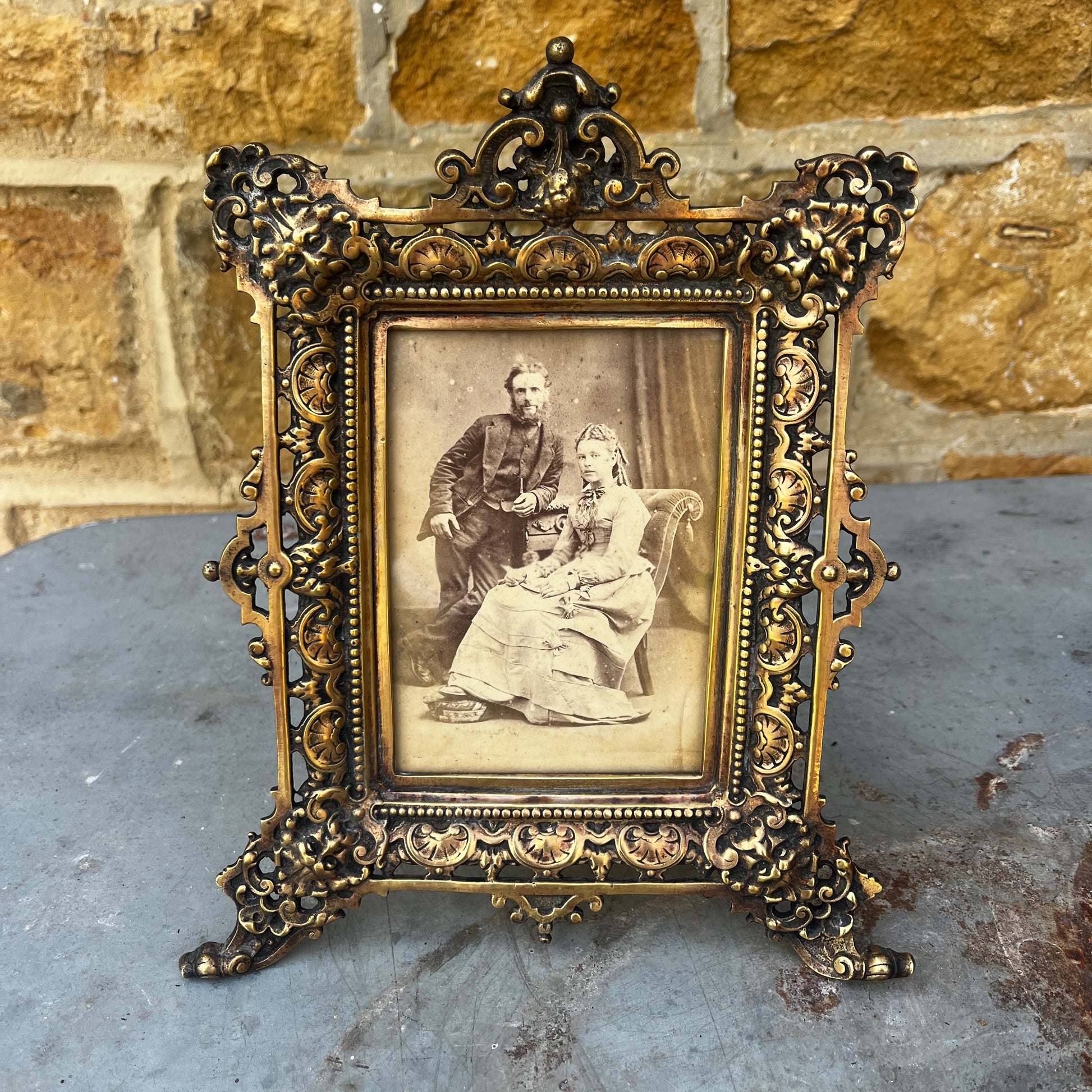 An ornate antique gilt metal photo frame with intricate pierced scrollwork, floral motifs, and a richly aged gold finish. The rectangular frame stands on decorative feet with an elaborate crest at the top, showcasing exceptional Victorian-era craftsmanship