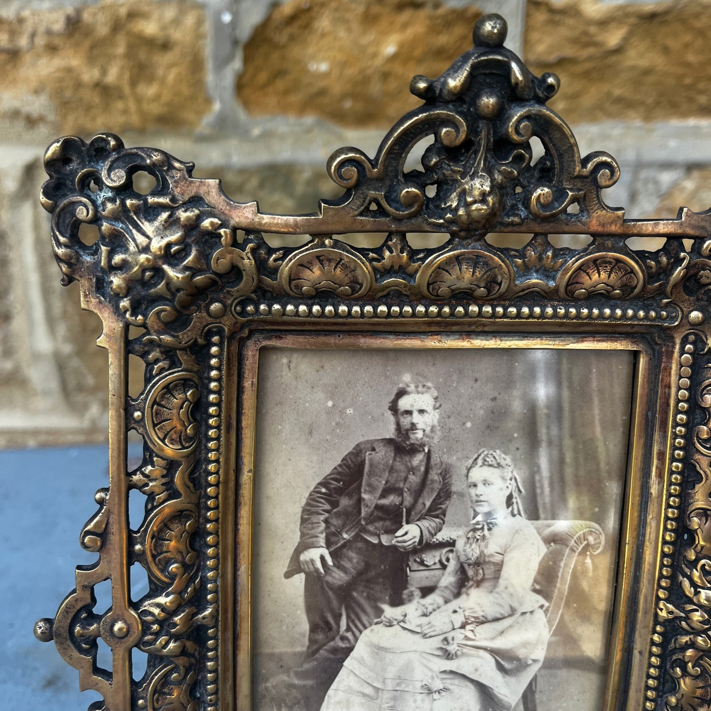 An ornate antique gilt metal photo frame with intricate pierced scrollwork, floral motifs, and a richly aged gold finish. The rectangular frame stands on decorative feet with an elaborate crest at the top, showcasing exceptional Victorian-era craftsmanship
