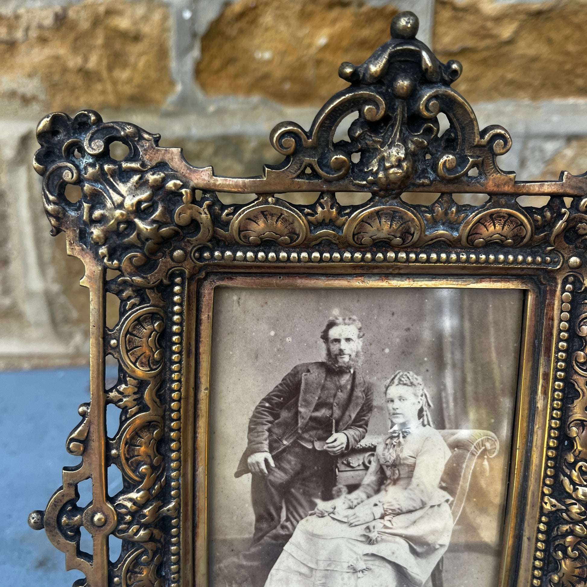 An ornate antique gilt metal photo frame with intricate pierced scrollwork, floral motifs, and a richly aged gold finish. The rectangular frame stands on decorative feet with an elaborate crest at the top, showcasing exceptional Victorian-era craftsmanship
