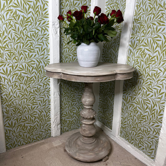 Small rustic wooden pedestal table with a clover-shaped tabletop - distressed in a stone colour.  The tabletop edge features carved contours with a small detail on the front edge.