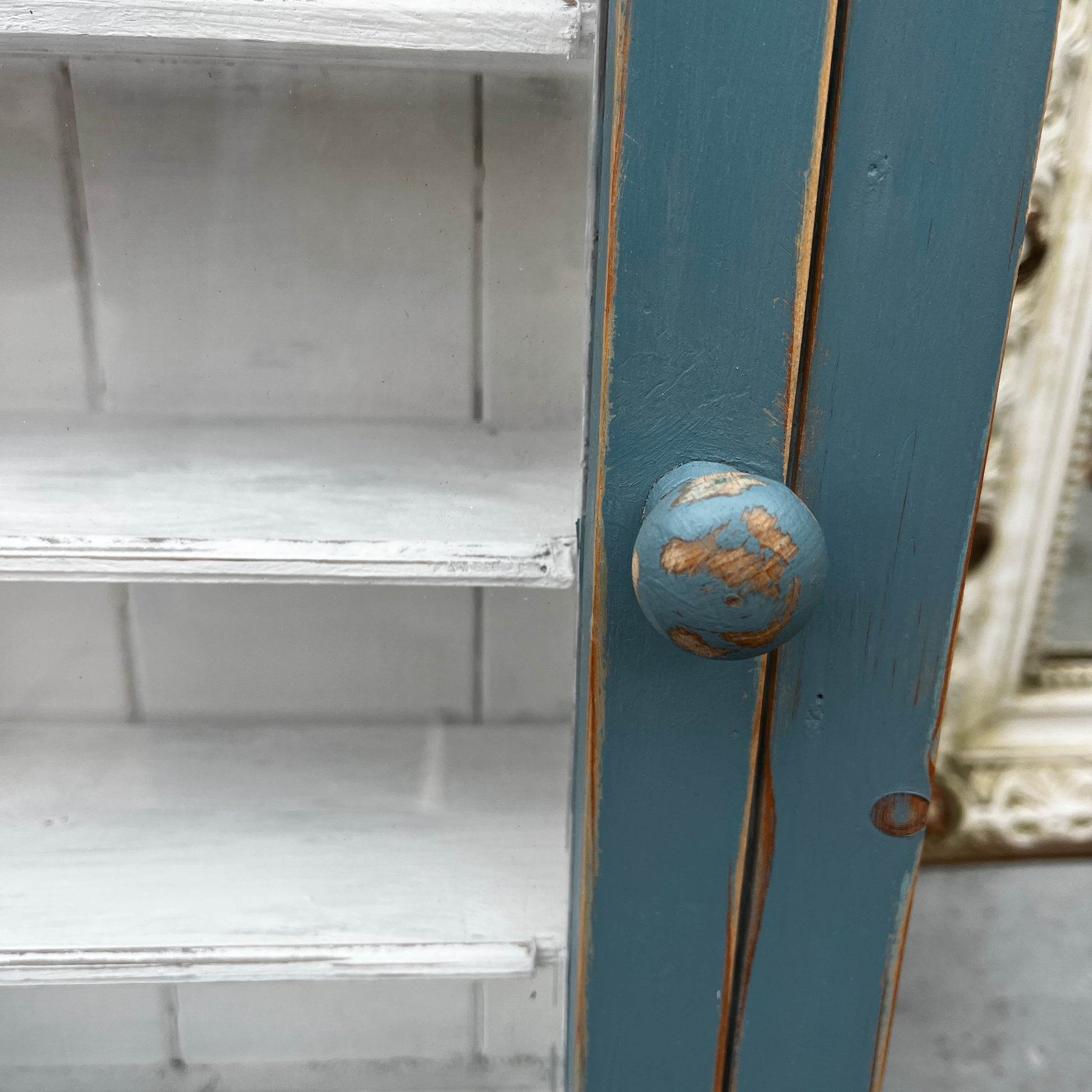 Vintage French wooden wall cupboard painted blue on the exterior and white inside, featuring a glass-front door that reveals internal shelves. The piece has a rustic, timeworn finish and classic country-style charm.