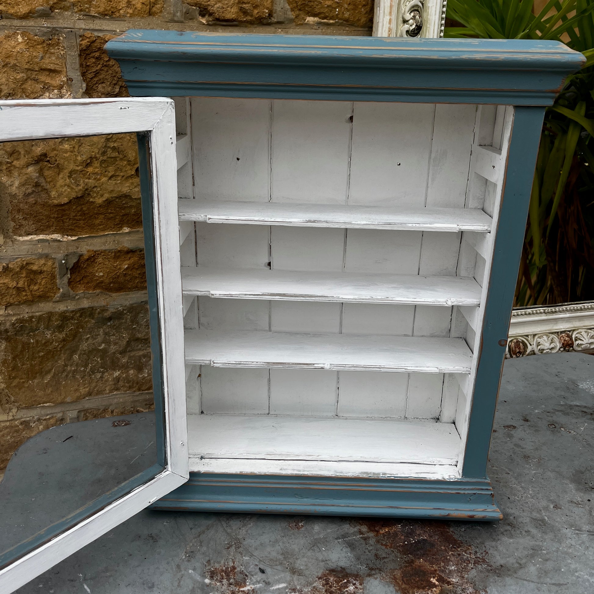 Vintage French wooden wall cupboard painted blue on the exterior and white inside, featuring a glass-front door that reveals internal shelves. The piece has a rustic, timeworn finish and classic country-style charm.