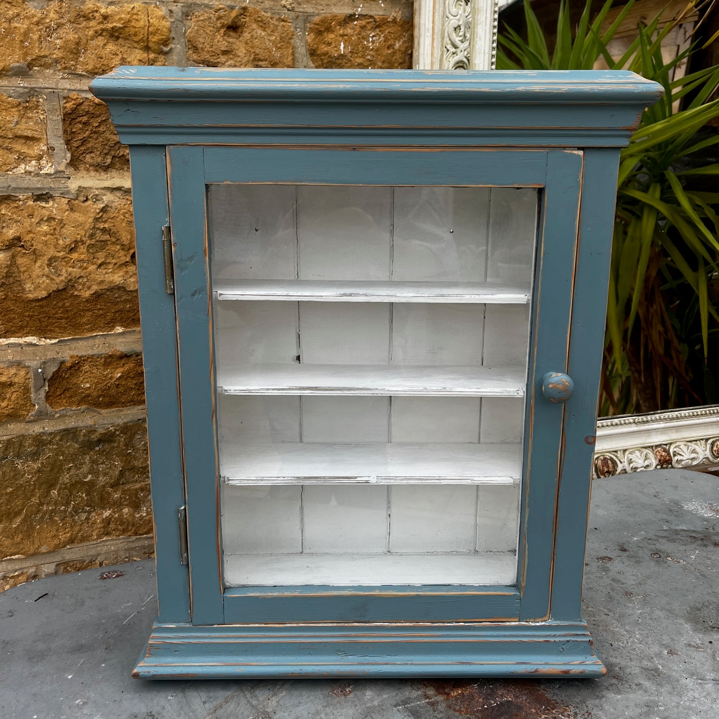 Vintage French wooden wall cupboard painted blue on the exterior and white inside, featuring a glass-front door that reveals internal shelves. The piece has a rustic, timeworn finish and classic country-style charm.
