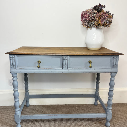 A French-style console table featuring a grey-blue distressed finish, with two shallow drawers adorned with small brass handles. The table stands on elegantly carved barley twist legs, exuding vintage charm