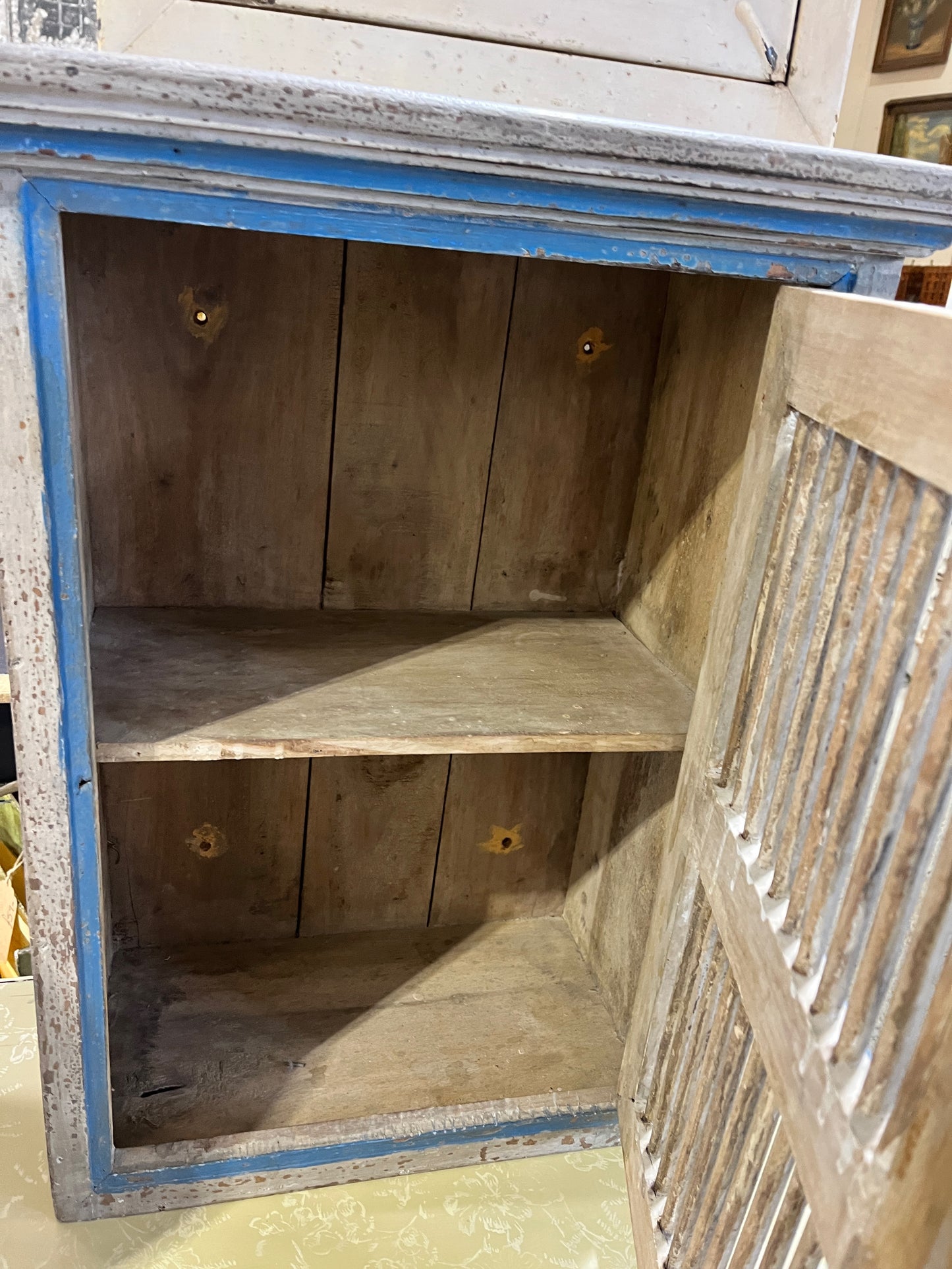 Small blue-grey Indian-style cupboard with a decorative door and one interior shelf, offering a blend of exotic charm and functional storage
