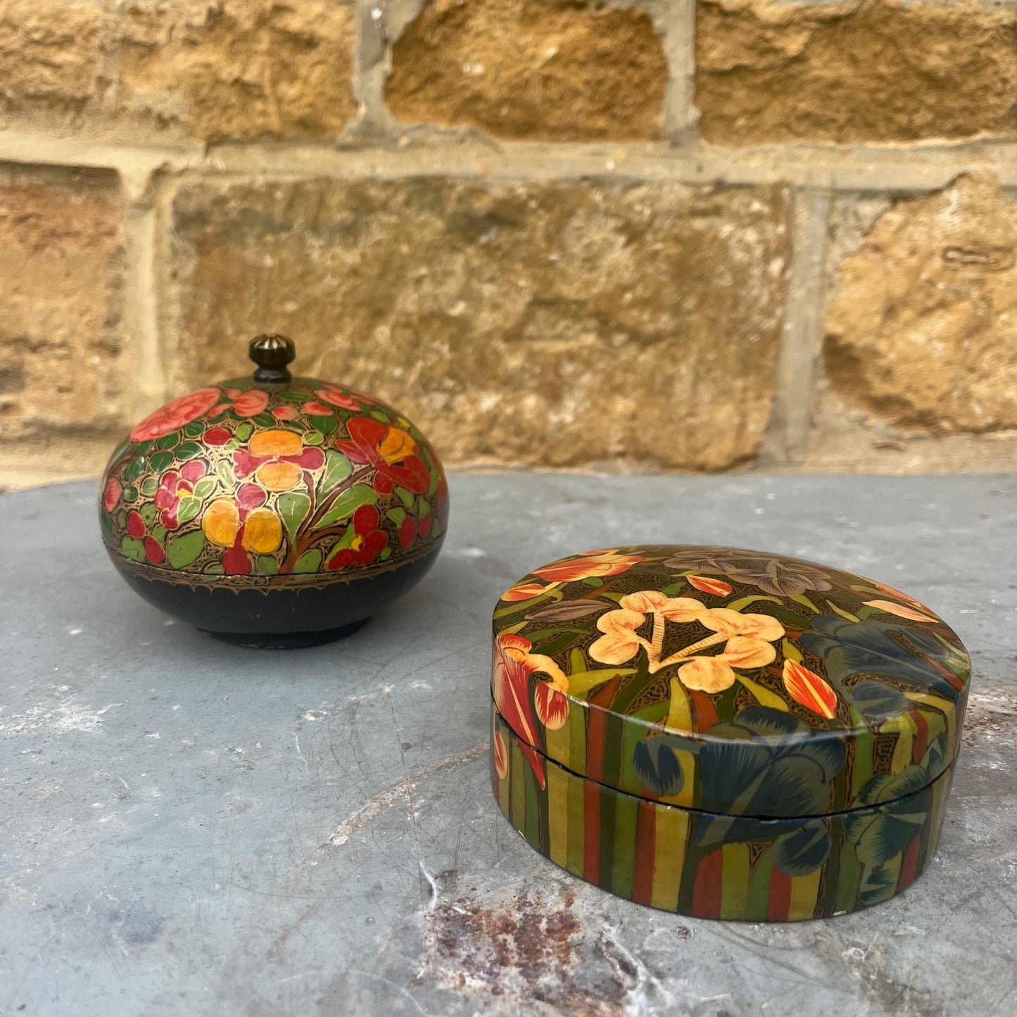 A decorative papier-mâché box with a fitted lid, adorned with vibrant, hand-painted floral patterns in shades of red, yellow, and green against a dark background.