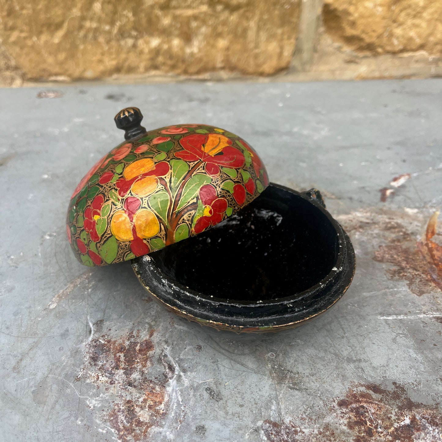 A decorative papier-mâché box with a fitted lid, adorned with vibrant, hand-painted floral patterns in shades of red, yellow, and green against a dark background.