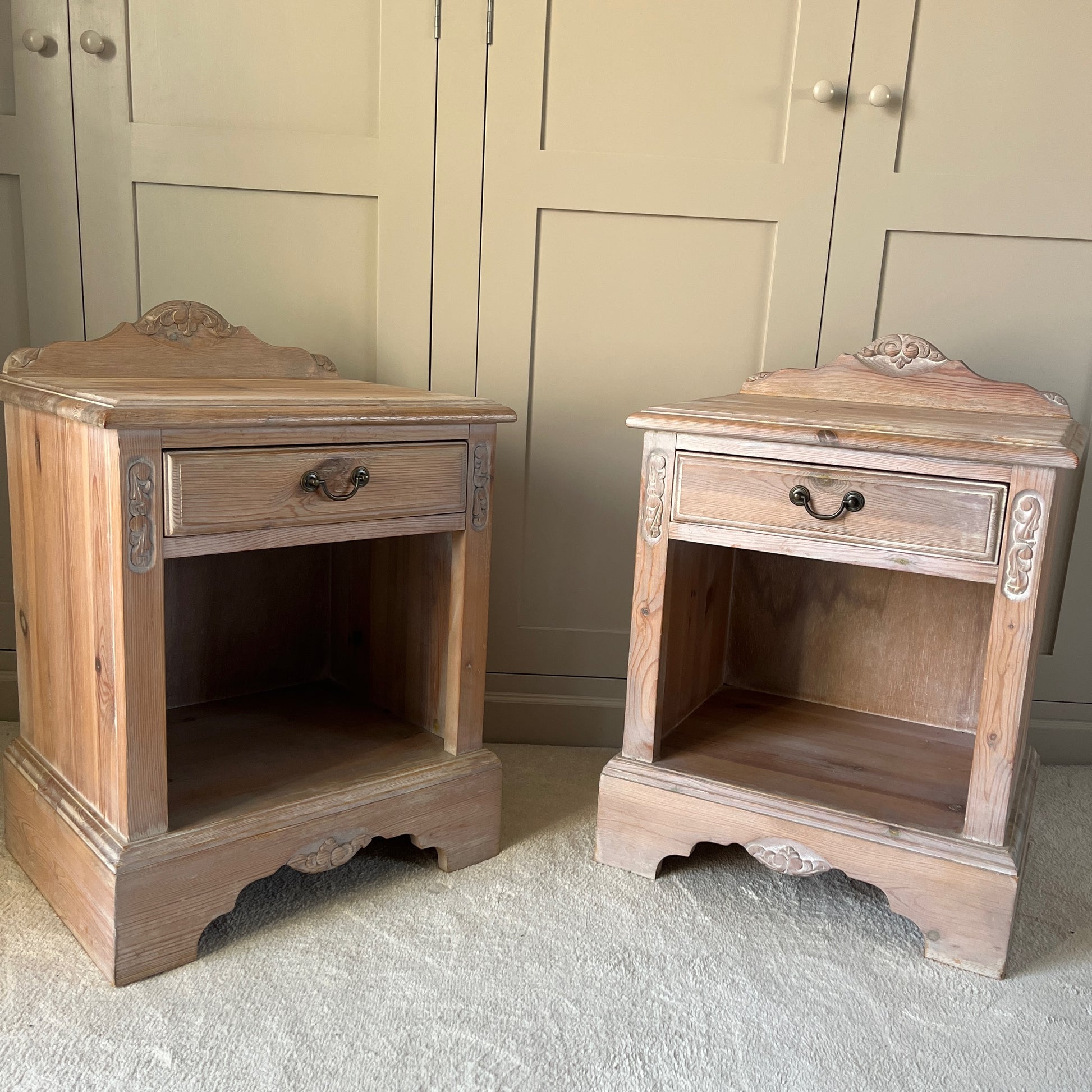 A pair of simple pine bedside tables with a natural wood finish, featuring decorative detail on the front, a single drawer with a ornate handle, and an open lower shelf, exuding rustic charm and functionality.