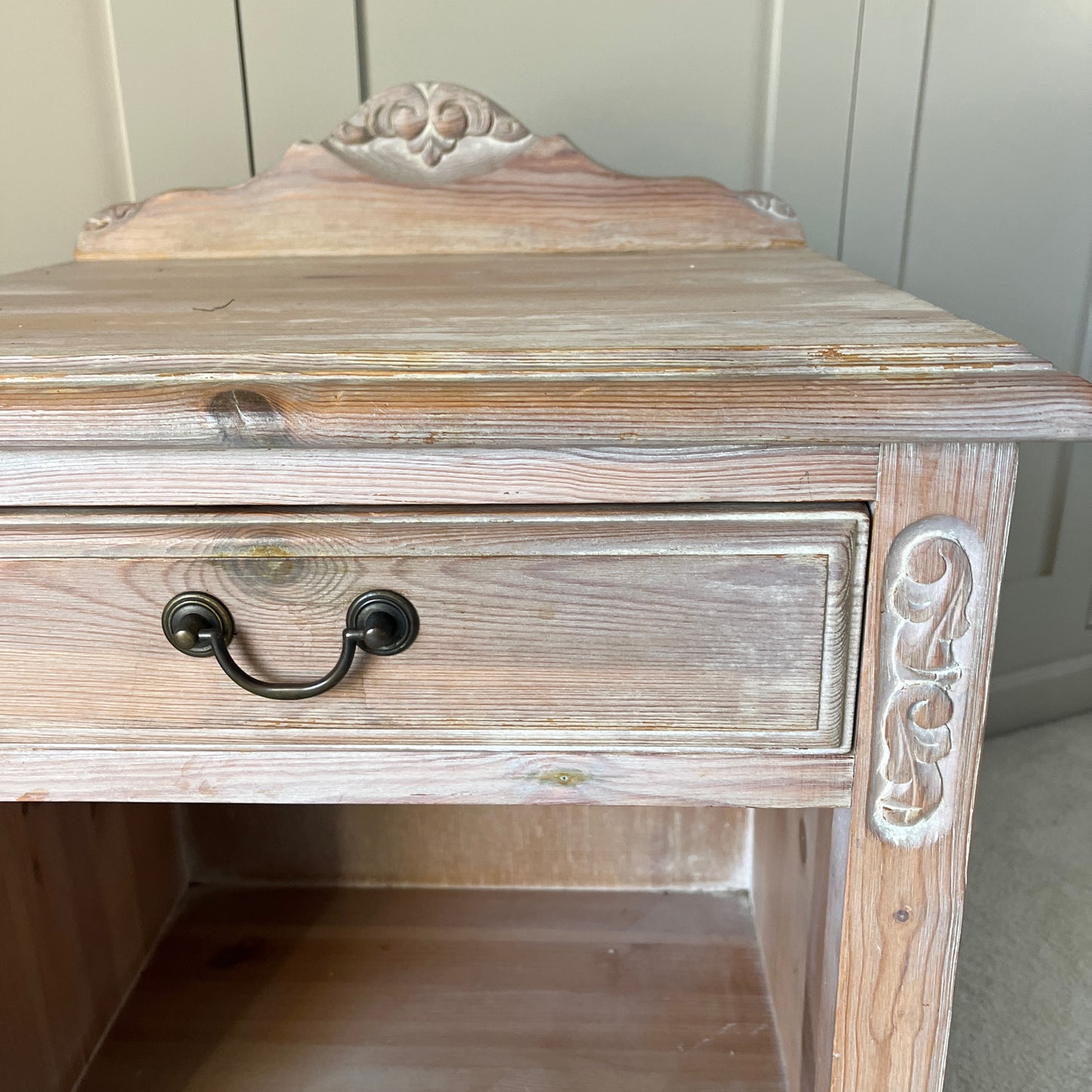 A pair of simple pine bedside tables with a natural wood finish, featuring decorative detail on the front, a single drawer with a ornate handle, and an open lower shelf, exuding rustic charm and functionality.