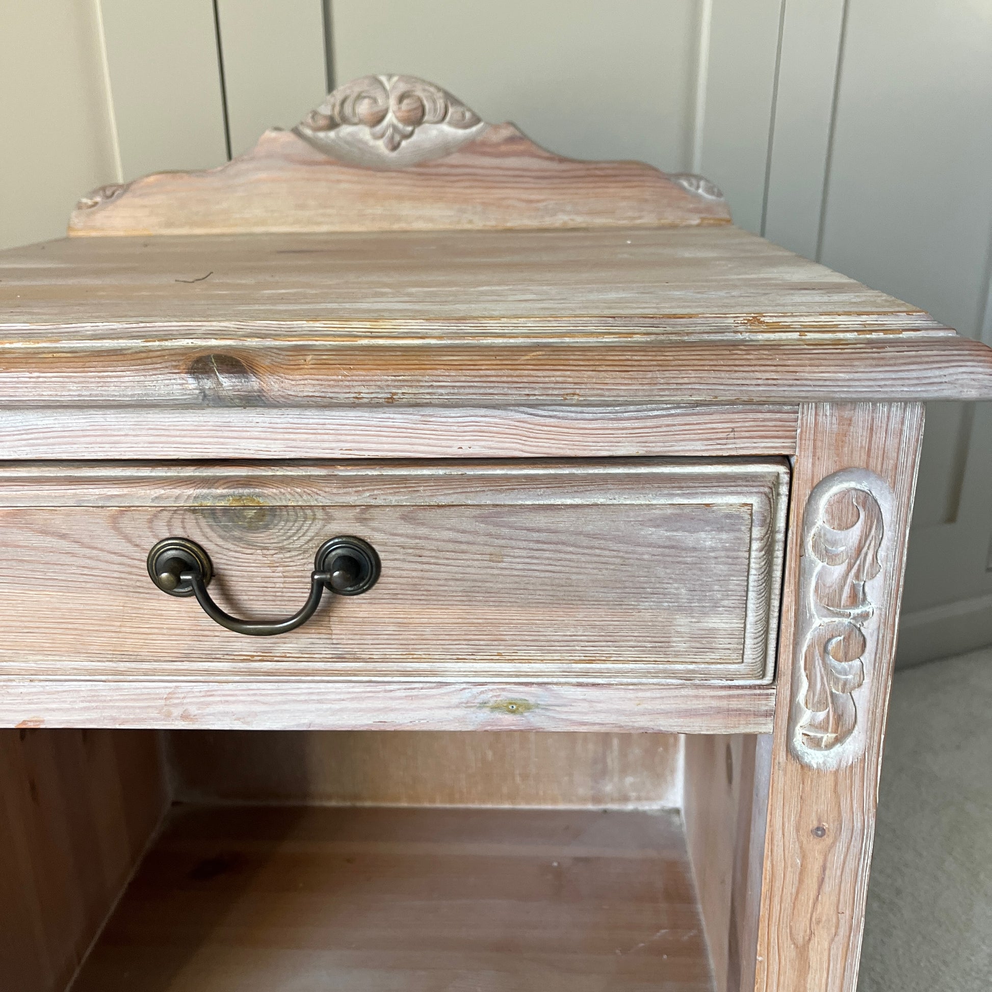 A pair of simple pine bedside tables with a natural wood finish, featuring decorative detail on the front, a single drawer with a ornate handle, and an open lower shelf, exuding rustic charm and functionality.