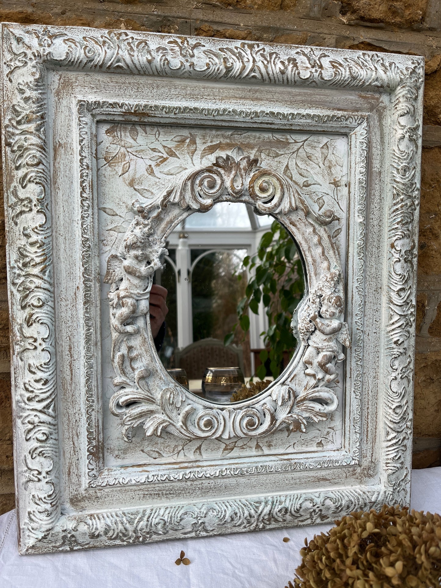 A shabby chic rectangular mirror with a distressed, white wooden frame, adorned with small, intricately carved angel figures positioned around the edge of the mirror. The vintage-style mirror reflects a soft, weathered charm.