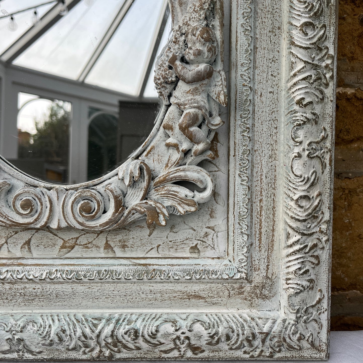 A shabby chic rectangular mirror with a distressed, white wooden frame, adorned with small, intricately carved angel figures positioned around the edge of the mirror. The vintage-style mirror reflects a soft, weathered charm.