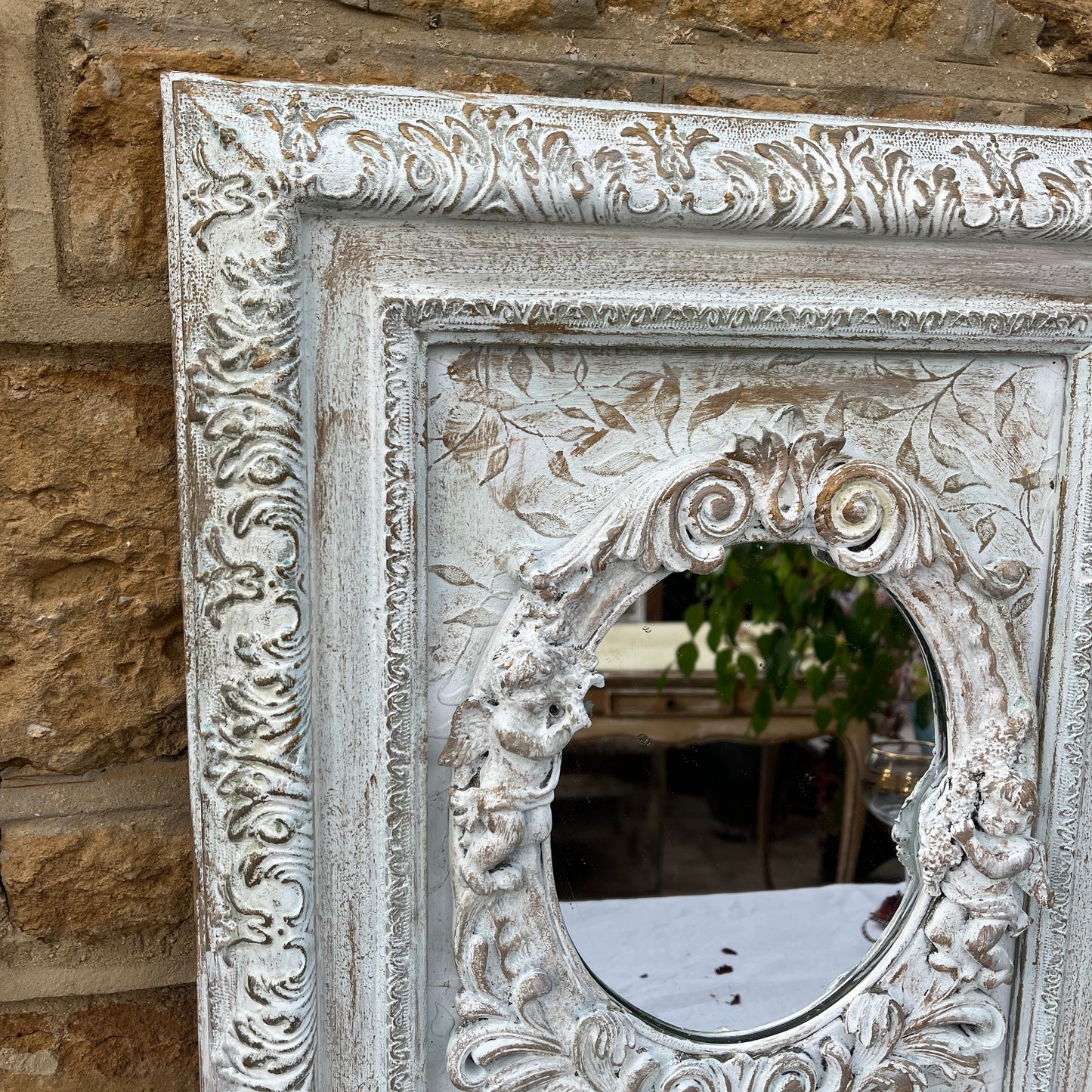A shabby chic rectangular mirror with a distressed, white wooden frame, adorned with small, intricately carved angel figures positioned around the edge of the mirror. The vintage-style mirror reflects a soft, weathered charm.