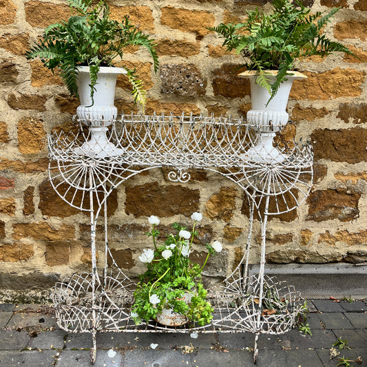 A Victorian decorative plant stand crafted from ornate wrought iron wire, featuring intricate scrollwork and floral patterns. The two-tiered stand, painted in a distressed white finish, supports small potted plants, exuding elegant vintage charm