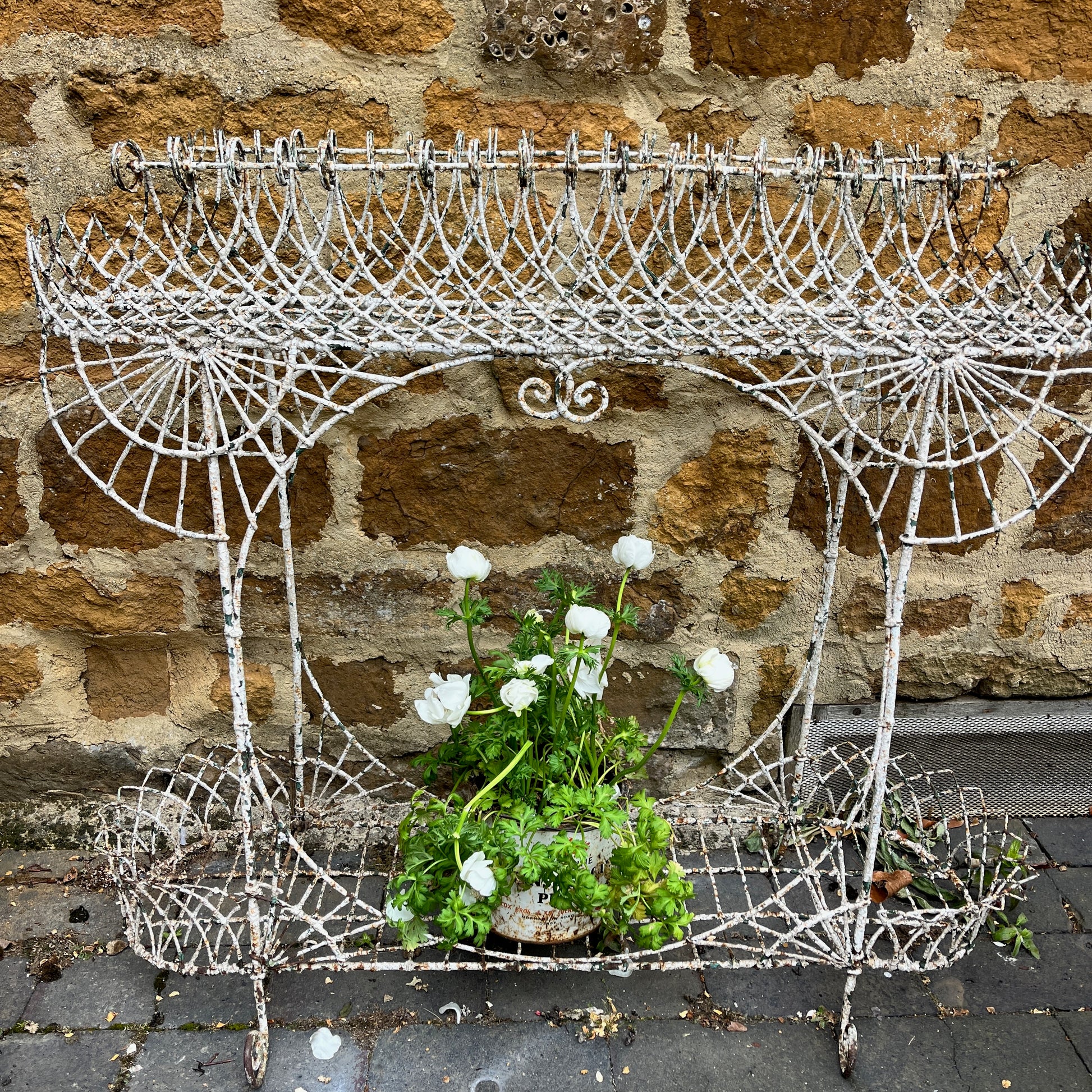 A Victorian decorative plant stand crafted from ornate wrought iron wire, featuring intricate scrollwork and floral patterns. The two-tiered stand, painted in a distressed white finish, supports small potted plants, exuding elegant vintage charm