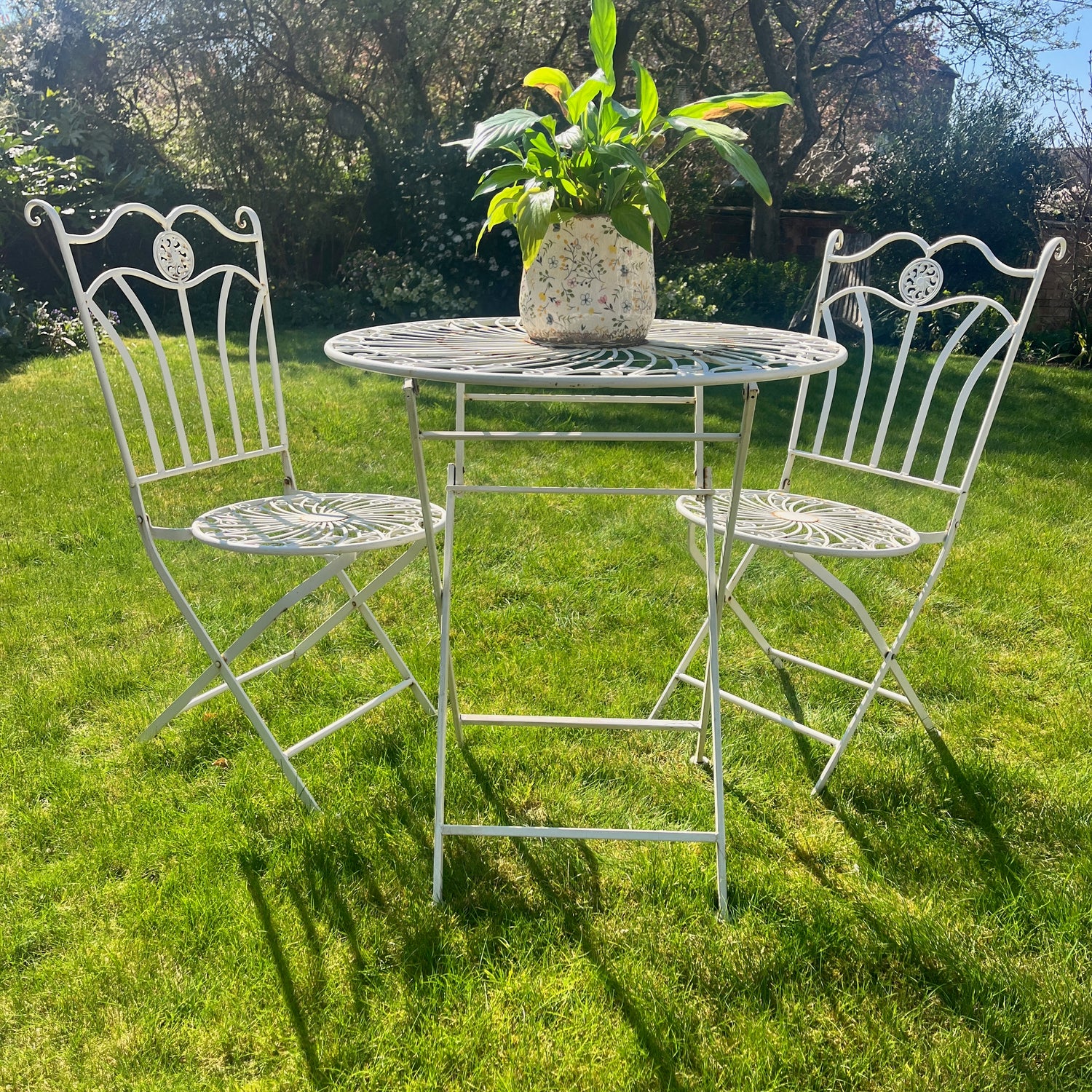 A decorative vintage bistro set crafted from white metal, including a small round table and two matching chairs, exuding a pretty and timeless outdoor charm
